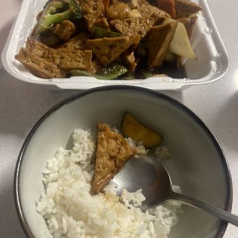 Shanghai Express - Fried tofu and vegetables - The image shows a styrofoam takeout container filled with stir-fried tofu, broccoli, carrots, and other vegetables in a sauce. In front, there is a bowl of white rice with a piece of tofu and a small piece of pumpkin on top. A metal spoon rests in the rice bowl. The setting appears to be on a light-colored table. - Chinese Near Me - Georgetown, Texas