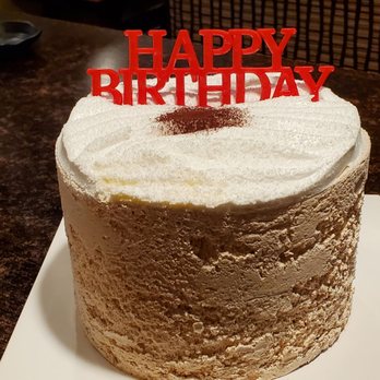 Tous Les Jours - Earl Grey Cake - A round birthday cake with white frosting and a textured surface, topped with a red "HAPPY BIRTHDAY" decoration and a dusting of cocoa powder in the center. The cake is placed on a white square base on a dark brown table. - Coffee & Tea Near Me - Midvale, Utah