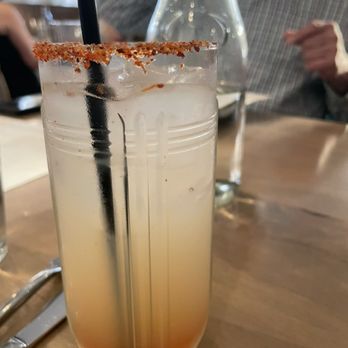 Lynette - Spicy boy - A tall, transparent glass contains a layered cocktail with orange and yellow colors, topped with a salted rim and a black straw. The drink is placed on a wooden table, with a glass bottle and a person in a checkered shirt visible in the background. The setting appears to be a restaurant or bar. - Beer Bar Near Me - Minneapolis, Minnesota