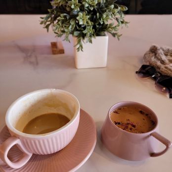 Cocomelt - The image shows two cups of hot beverages on a table, one with a greenish drink in a pink cup with a matching saucer, and the other with a darker liquid in a pink mug. A potted plant with green foliage is positioned behind the cups, and sunglasses along with a cloth are on the right side of the table. The background features a colorful, illuminated decorative element, possibly a fireplace or light display. - Desserts Near Me - Lombard, Illinois