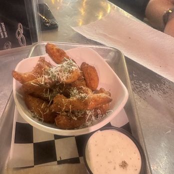 The Dive - Cheese Curds - A serving of potato wedges seasoned with spices and topped with shredded cheese and herbs is presented in a white bowl on a metal tray. A small cup of creamy dipping sauce is placed beside the bowl. The tray is on a table with a paper liner, and the background shows a restaurant setting with chairs and a person partially visible. - Pop-Up Restaurants Near Me - Sioux Falls, South Dakota