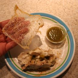 One of these things does not belong in a burrito. - A plate holds cooked chicken, a small cup of green sauce, and a hand holding a piece of raw fish or seafood.