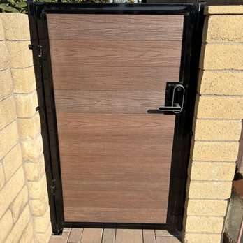 Socal Gates - A wooden gate with a black metal frame and handle is mounted between two brick walls, leading to an outdoor area. - Fences & Gates Near Me - Orange, California