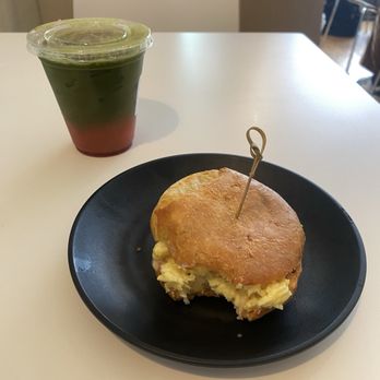 Bōm Dough - fiz matcha, strawberry, ginger sunny stack - A breakfast sandwich with scrambled eggs and bread on a black plate, accompanied by a layered matcha latte with red bean on a white table. - Brazilian Near Me - Cambridge, Massachusetts
