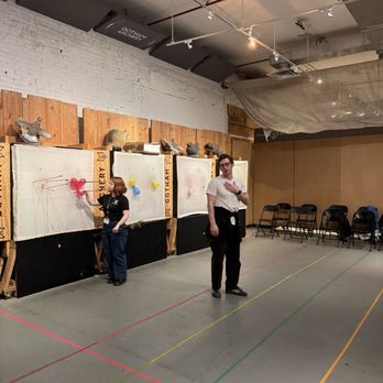 Gotham Archery - Instruction that's not just easy but insanely fun! - The image shows an indoor archery range at Gotham Archery in Brooklyn, NY. Two individuals are present: one woman is aiming a bow at targets on a wall, while a man stands nearby with his hand on his chest. The range features a white brick wall, wooden target backings with mounted animal heads, and a row of chairs along the side. The ceiling has exposed pipes and lighting fixtures, and the floor is marked with colored lines for shooting lanes. - Archery Near Me - Brooklyn, New York
