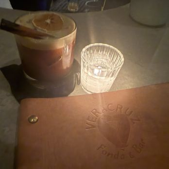 La Mezca - A cocktail featuring a cinnamon stick and orange slice garnish is positioned on a table, accompanied by a clear glass of water. The table surface appears to be made of stone or similar material. A leather menu or book with embossed text and a heart logo is partially visible in the foreground. The setting suggests a bar or restaurant environment with dim lighting. - Cocktail Bars Near Me - Austin, Texas