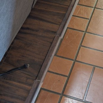 Floor Daddy - They installed a small piece of transition strip that's not even the same kind as the other.. - The image shows a transition strip installed between a wood floor and a tile floor. The transition strip is made of wood and appears to be slightly misaligned or not matching the adjacent flooring materials. A black cable is lying on the wood surface near the transition strip. The photo captures the edge where the two flooring types meet, with visible grout lines on the tile and wood grain on the wood flooring. - Flooring Near Me - Tempe, Arizona
