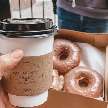 Underground Donut Tour - Doughnut Vault - A person is holding a white paper coffee cup with a black plastic lid, wrapped with a brown sleeve labeled 'Doughnut Vault Old-Fashioned Chicago.' In the background, there is a cardboard box containing several glazed donuts with powdered sugar, placed on a wooden surface. Part of a person wearing a gray jacket and blue jeans is visible behind the table, with a metal railing and outdoor scenery beyond. - Food Tours Near Me - Chicago, Illinois