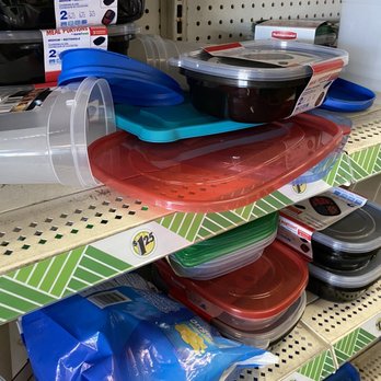 Dollar Tree - Mess. - Multiple plastic food storage containers in colors such as red, black, blue, and green are stacked on a store shelf. Some containers have clear lids, and packaging labels are visible on some items. The shelf has a green and white patterned border, and other food storage products are seen nearby, indicating a retail environment. - Discount Store Near Me - Pompano Beach, Florida