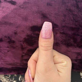 Charming Nails - The image features a hand with a pink glitter nail polish on the thumb, with the other fingers partially visible. The background consists of a purple fabric with a velvety texture. The focus is on the well-manicured nails, highlighting the glittery finish and the neat appearance of the manicure. - Nail Salons Near Me - Washington, DC, District of Columbia