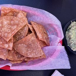 Guacamole - A basket filled with fried tortilla chips, lined with pink paper, is placed on a dark surface. Next to it, there is a small black bowl containing guacamole with visible chunks of avocado and other ingredients. The setting suggests a casual dining or snack presentation, typical of Mexican cuisine.