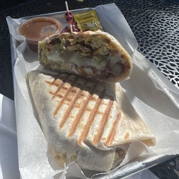 Cafe Urbano - Breakfast Burrito (D) - added bacon - A breakfast burrito with grill marks is placed on a metal tray lined with parchment paper. The burrito is sliced open, revealing eggs, bacon, and vegetables inside. A small container of sauce and a packet of condiments are also on the tray. The setting appears to be outdoors, with sunlight casting shadows on the tray. - Cafes Near Me - Chicago, Illinois