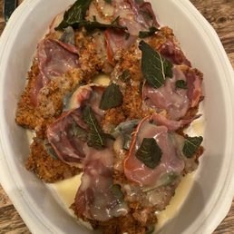 A serving dish contains breaded meat cutlets topped with slices of prosciutto, fresh sage leaves, and melted cheese. The dish is placed on a wooden table, with utensils visible in the background. The presentation includes herbs and a creamy sauce around the meat.