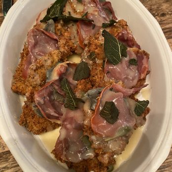 Chef Christopher Hensel - A serving dish contains breaded meat cutlets topped with slices of prosciutto, fresh sage leaves, and melted cheese. The dish is placed on a wooden table, with utensils visible in the background. The presentation includes herbs and a creamy sauce around the meat. - Personal Chefs Near Me - Sag Harbor, New York