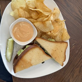 Jules' Bistro - My $20 sandwich. It only had avocado mushrooms and sauerkraut.  I opted not to pay even more for vegan cheese - A white square plate contains a serving of potato chips, a small bowl of creamy dipping sauce, a pickle spear, and a toasted sandwich with lettuce and meat. The plate is placed on a wooden table, with part of a black cloth or napkin visible at the top of the image. The sandwich is cut diagonally, showing the filling inside. The chips are piled on one side of the plate, and the dipping sauce is in a small round bowl. - New American Near Me - St. Cloud, Minnesota