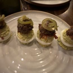 Four appetizer bites are arranged on a white plate, each consisting of a hard-boiled egg half topped with seasoned ground meat, crispy bacon, and a slice of jalapeño pepper. The plate is set on a dark wooden table with a black napkin and additional plates in the background. The image appears to be taken in a restaurant setting.