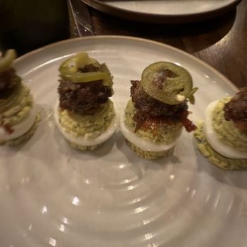Yard House - Four appetizer bites are arranged on a white plate, each consisting of a hard-boiled egg half topped with seasoned ground meat, crispy bacon, and a slice of jalapeño pepper. The plate is set on a dark wooden table with a black napkin and additional plates in the background. The image appears to be taken in a restaurant setting. - New American Near Me - Washington, DC, District of Columbia