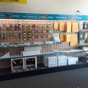 The UPS Store - The image shows a retail shelving unit with packaging supplies, such as tape, boxes, and wrapping paper, arranged on a pegboard wall. Several boxes and items are missing or not fully stocked, and some packaging materials are stacked or placed on the shelves. The store has a sign indicating it is a packing store, with a yellow sign and a blue banner. The floor is carpeted, and the background includes a plain wall with ceiling lights. - Shipping Centers Near Me - Mesa, Arizona
