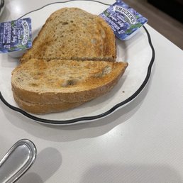 Rye bread dry - Two slices of toasted bread are arranged on a white plate with a black rim, with one slice slightly overlapping the other. The bread appears toasted with a golden-brown crust and some uneven toasting marks. Two butter packets labeled 'Smucker's' are positioned at the top corners of the plate. The plate is on a white surface, and part of a metal utensil is visible in the lower left corner. The background includes a glimpse of a dark floor and furniture.