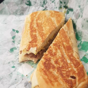 Cafe Cubano - Classic Cuban Club (Pressed Sandwich) - A pressed Cuban sandwich, cut in half, showing layers of ham, roasted pork, Swiss cheese, and pickles inside a toasted Cuban bread. The sandwich is placed on green and white patterned paper, with a hand holding the paper in the background. The image emphasizes the sandwich's golden-brown crust and filling. - Cuban Near Me - Springfield, Missouri
