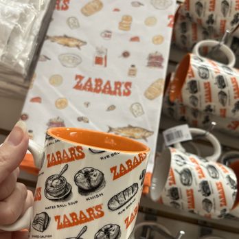 Zabar's - The perfect gift - A ceramic mug with Zabar's branding and food illustrations is held in front of a display of matching mugs at Zabar's store. - Bakeries Near Me - New York, New York