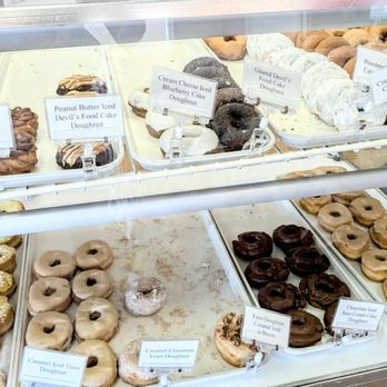 Henze's Bakery - The image shows a glass display case filled with a variety of donuts and baked goods at Henze's Bakery in Valparaiso, IN. The case contains multiple trays with different types of donuts, including chocolate iced, caramel cinnamon, glazed devil's food cake, and others, each labeled with small signs. The baked goods are arranged in neat rows, and the labels provide details about each item. The bakery has an old-fashioned atmosphere, emphasizing traditional recipes and a family-owned history. - Bakeries Near Me - Valparaiso, Indiana