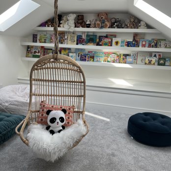 Dream House Services - Custom Shelving - A cozy children's reading nook includes a hanging wicker chair with a panda plush toy, a decorative pillow, and a fluffy blanket. Behind, a white bookshelf holds children's books and stuffed animals, with skylights providing natural light. The space has a soft gray carpet and a dark round cushion on the floor. - General Contractors Near Me - Randolph, Massachusetts