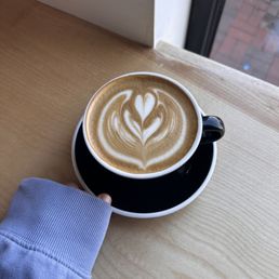 Oat Milk Latte - A latte with detailed foam art in a black ceramic cup is placed on a matching black saucer on a light wooden table. A person's hand, wearing a light-colored sleeve, is holding the cup. The setting is near a window, with visible brick pavement outside. The image captures the coffee's presentation and the natural lighting from the window.