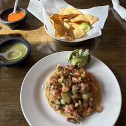 Tostada - A plate holds a tostada topped with diced tomatoes, onions, cilantro, and avocado slices, garnished with a small lime wedge. Two small bowls contain sauces—one red and spicy, the other green and creamy. The dish is served on a wooden table with a basket of tortilla chips and additional sauces nearby, in a casual dining setting.