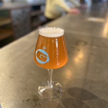 Gravity Bound Brewing - Lemon Layer - A tulip-shaped beer glass with a logo on it contains an amber-colored beer with a foamy head. The glass is placed on a concrete bar counter, with a blurred background showing a person in a yellow shirt and bar elements, indicating an indoor bar or brewery environment. - Brewpubs Near Me - Albuquerque, New Mexico