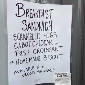 Bread & Roses - Breakfast sandwich menu - A handwritten menu sign on a glass surface lists breakfast sandwich options, including scrambled eggs, Cabot cheddar cheese, and choices of fresh croissant or homemade biscuit. The sign mentions availability with veggie sausage and notes the option of a breakfast burrito with scrambled eggs, cheddar, and other ingredients. The sign is written in black marker on white paper, with a smaller note attached at the bottom providing additional details. - Coffee & Tea Near Me - Wells, Maine