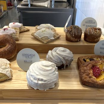 Window Coffee Bar - Pastries - A variety of pastries including a cinnamon roll, lemon scones, and fruit Danish are arranged on a wooden display tray behind a glass window. The pastries are labeled with small signs indicating their names and prices, such as cinnamon roll for $5.75 and fruit Danish for $7.50. The display is set in a bakery or coffee shop environment with a stainless steel counter and kitchen equipment visible in the background. - Coffee & Tea Near Me - Phoenix, Arizona