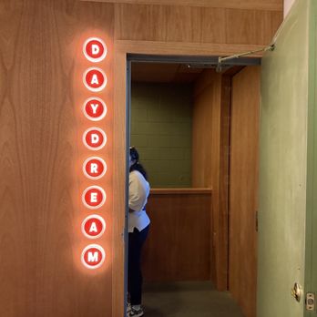 Daydream - Hidden entrance - A wooden door is partially open, revealing a person standing inside with their back turned. To the left of the door, a vertical sign with illuminated red circular letters spells "DAYDREAM." The sign is mounted on the wooden wall, and the interior visible through the open door has green tiled walls. A hand is visible holding the green door handle on the right side of the image. - Cocktail Bars Near Me - Albuquerque, New Mexico