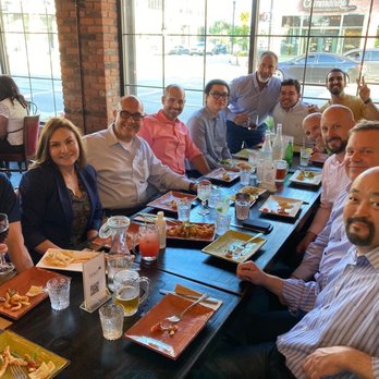 Mint 29 - Team dinner - A group of twelve people seated at a long table in a restaurant with large windows, enjoying a meal together during a team dinner. - Bars Near Me - Dearborn, Michigan