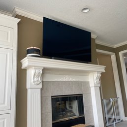 TV installation - A large flat-screen TV is mounted above a white, ornate fireplace mantel in a living room with beige walls and crown molding.