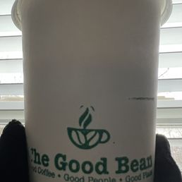 Cinnamon French Toast Latte - A white disposable cup with The Good Bean logo and slogan, held by a person wearing a black glove, with window blinds in the background.