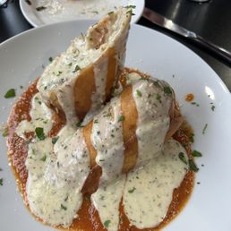 The Kevin Costner - A dish featuring two stuffed pastry items topped with a white creamy sauce, garnished with herbs, served on a white plate.