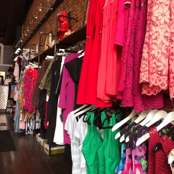 Blush On Main - Inside a boutique clothing store, multiple racks hold a variety of dresses and apparel in vibrant colors and patterns. The display includes red, pink, green, and multicolored garments, with some lace and patterned fabrics. The store has an exposed brick wall and overhead lighting, creating a modern, stylish shopping environment. - Women's Clothing Near Me - Madison, Indiana