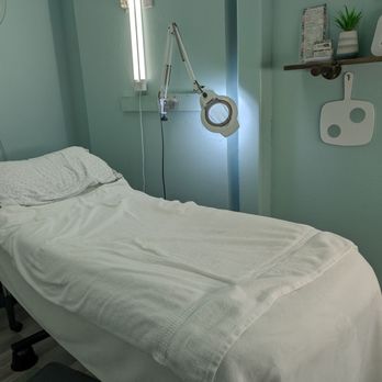 Mama Bare Sugaring Studio - Ormond Beach - Comfortable beds - A treatment bed covered with white linens is in a room with mint green walls, featuring a magnifying lamp and decorative items on a shelf. - Sugaring Near Me - Ormond Beach, Florida