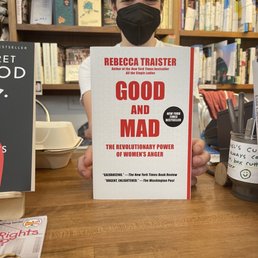 A woman with dark hair and a black face mask displays the book 'Good and Mad' by Rebecca Traister in a bookstore setting with shelves of books in the background.