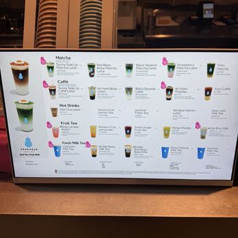 Uluhuluh - The illuminated digital menu board lists a variety of beverages such as matcha lattes, fruit teas, and milk teas, with images and descriptions. The menu includes options like Sunny Side Up Matcha Latte, Red Bean Matcha Latte, and Yuzu Matcha, among others. The background shows part of the cafe interior, with stacked cups and a counter area. - Bubble Tea Near Me - Brooklyn, New York