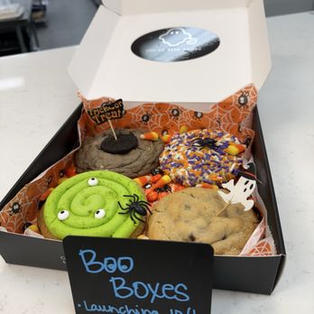 Half Baked Goodness - A box of decorated cookies with Halloween themes, including a green spiral cookie with eyes, a chocolate cookie with a spider, and a cookie with purple and orange sprinkles, on a white surface. - Desserts Near Me - Naperville, Illinois