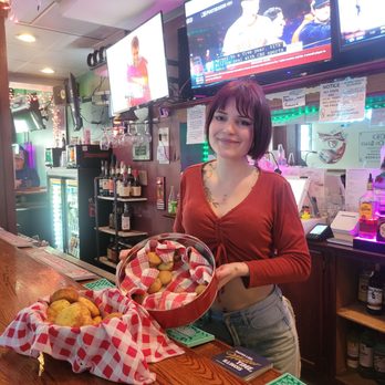 Stella's On State - A woman with short dark hair and a red top stands behind a bar counter holding a basket of baked goods, with more baked items on the counter. The background features multiple TV screens and bar supplies. - Dive Bars Near Me - Lockport, Illinois