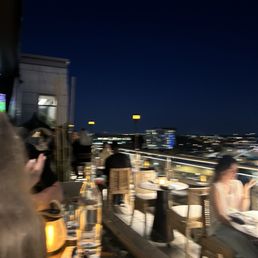 An outdoor rooftop dining space at night with several patrons seated at tables. The city skyline is visible in the background with illuminated buildings and streetlights. Some people are engaged with their phones, and there are bottles and glasses on the tables. The scene includes warm lighting and a railing along the edge of the rooftop, providing a view of the cityscape.