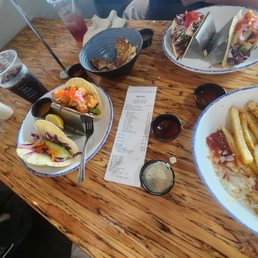 A wooden table holds tacos, fries, a slice of pie, drinks, and a receipt, indicating a casual dining setting with American and fast-food items.