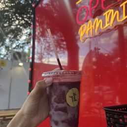 A hand with a tattoo holds a berry smoothie in a clear plastic cup with a lid and straw, positioned in front of a red food truck. The food truck features neon signs that read 'Open' and 'Panini,' and the background includes trees and outdoor seating. The scene suggests an outdoor food truck setting with fresh ingredients and vibrant signage.