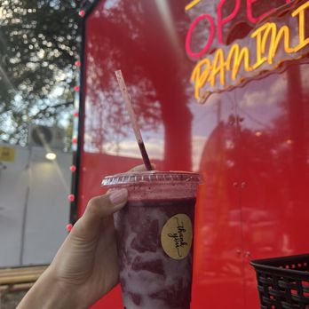 Paninik - A hand with a tattoo holds a berry smoothie in a clear plastic cup with a lid and straw, positioned in front of a red food truck. The food truck features neon signs that read 'Open' and 'Panini,' and the background includes trees and outdoor seating. The scene suggests an outdoor food truck setting with fresh ingredients and vibrant signage. - Food Trucks Near Me - Austin, Texas