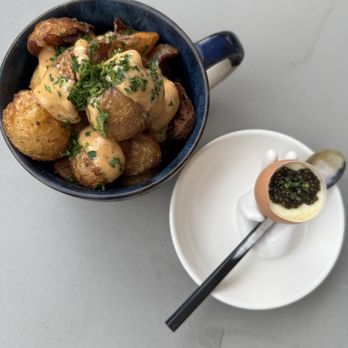 I Egg You - Umami Egg, Basic Potatoes - A mug filled with roasted potatoes garnished with herbs, accompanied by a soft-boiled egg topped with caviar on a white plate with a spoon. - Breakfast & Brunch Near Me - Washington, DC, District of Columbia