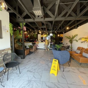 787 Coffee - The image shows the interior of a cafe with a mix of seating arrangements, including chairs and sofas, and decorative plants along the walls. The ceiling is painted black with exposed beams and ductwork. A yellow caution sign is placed on the floor, indicating a wet floor. The bar area in the background has a counter with a person standing nearby, and string lights and a neon sign add ambiance to the space. - Coffee & Tea Near Me - El Paso, Texas