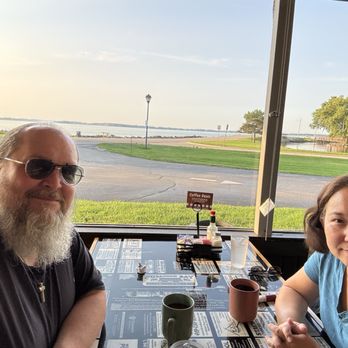 The Bay - The view of the lake from our table - A man with a beard, sunglasses, and a cross necklace sits at a table with a woman inside a restaurant featuring large windows. The view outside shows a lake, grassy areas, trees, and a clear sky. The table has cups, condiments, and menus, indicating a dining setting near water. - American Near Me - Celina, Ohio